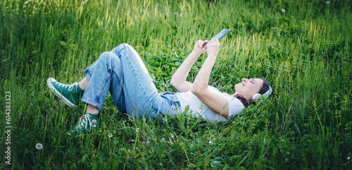 Fototapeta Young cheerful woman in a white t-shirt lying on the green grass in the garden looking into the smartphone screen