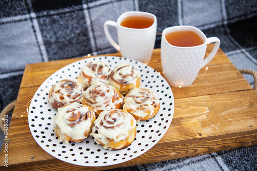 Two cups of black tea stand on a wooden tray on the sofa with a black and white checkered plaid. Fresh and fragrant cinnamon rolls close up lie on a plate with polka dots  beautiful morning