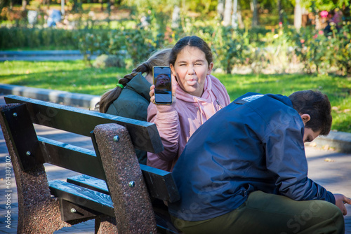 Children are sitting on a bench in the park. One girl with a phone turned around and teases.