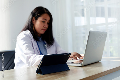 Asian female doctor working in hospital office On the table there is a clipboard and a computer. focus on laptop