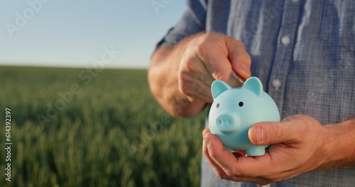 A man puts coins in a piggy bank, stands against the background of a field where wheat grows