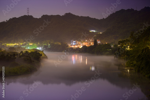 Night fog floats and moves along the river. Hazy and dreamy scenery. The colorful lights of the village reflect on the river.