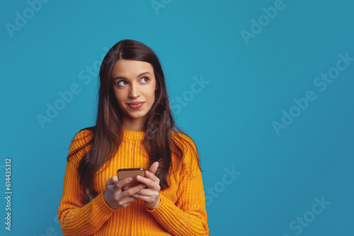 Dreamy young good looking girl holds mobile phone has thoughtful expression thinks about received message looks away isolated over blue background