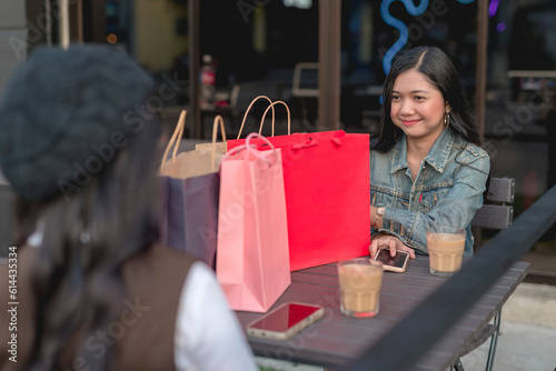 A pretty lady in a denim jacket smiles at a lady friend sitting in front of her at a restaurant. At their table are their shopping bags, coffees and phones. A window glass in the background