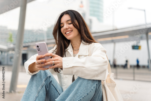 Bright and cheerful young woman with shopper on shoulder waiting public transport at station smiling beautifully while texting on phone