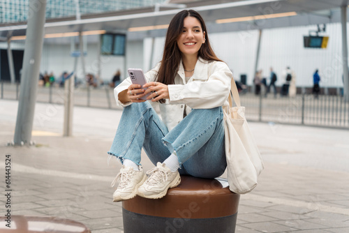 Bright portrait of cheerful young woman 25s with shopper on shoulder waiting public transport at station smiling beautifully and texting