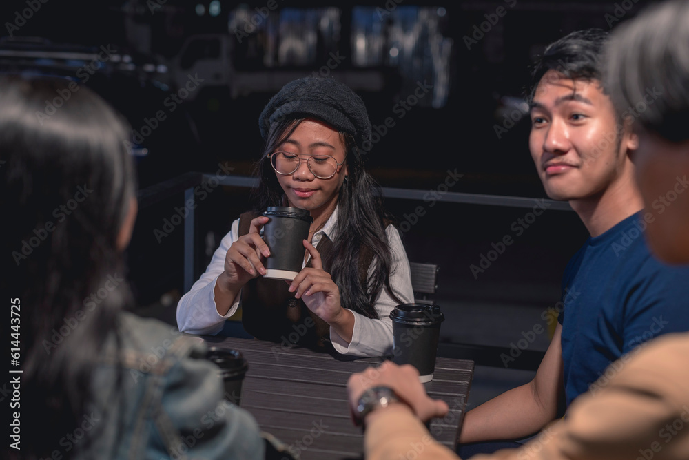 A pretty lady in beret, glasses and brown sweater vest is looking intensely at her coffee, holds it with both hands while her other friends are conversing. A truck, rail and dark in the background.