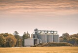 Steel grain silos stand next to a field. Agro-Industrial landscape. Steel Silos Embrace Vast Fields, Serving as Epitomes of Agricultural Storage and Processing