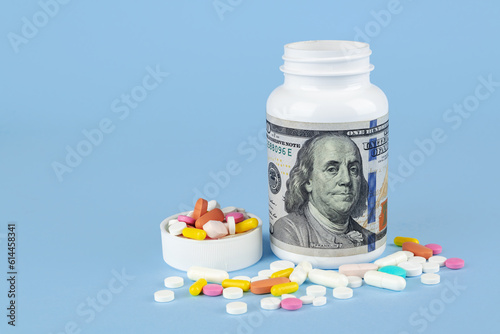 Pills spilling out of pill bottle with dollar banknote on blue background. Expensive medicine concept.