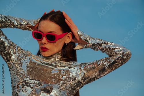 Fashionable confident woman wearing trendy fuchsia color rectangular sunglasses, sequin  turtleneck top, posing outdoor, against blue sky. Close up fashion portrait. Copy, empty space for text