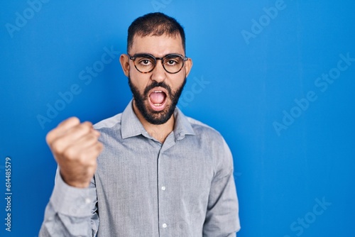 Middle east man with beard standing over blue background angry and mad raising fist frustrated and furious while shouting with anger. rage and aggressive concept.