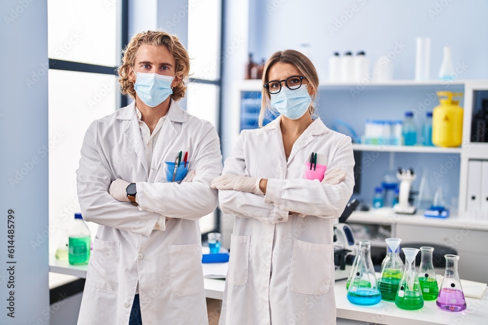 Man and woman wearing scientist uniform and medical mask standing with arms crossed gesture at laboratory Man and woman wearing scientist uniform and medical mask standing with arms crossed gesture at laboratory