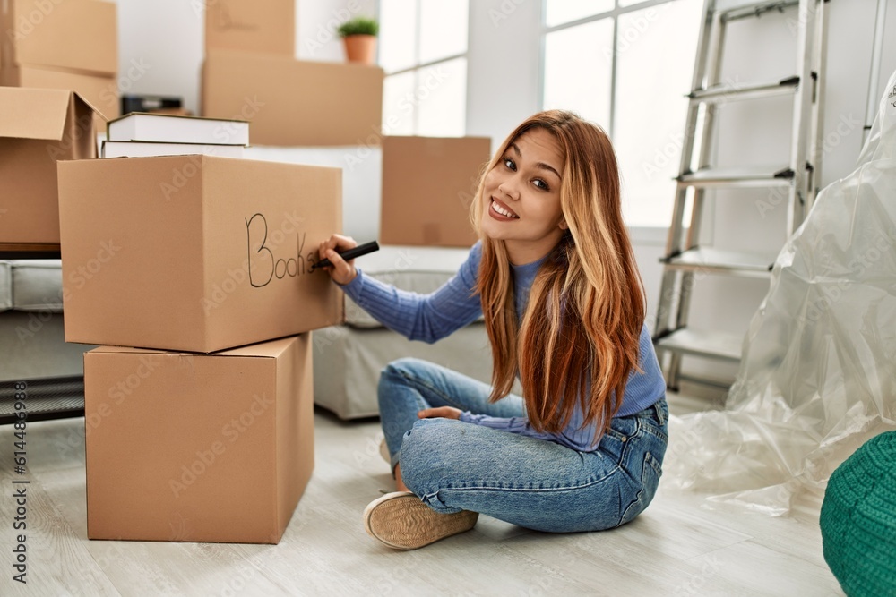 Young caucasian woman smiling confident writing books word on cardboard box at new home Young caucasian woman smiling confident writing books word on cardboard box at new home