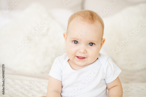 portrait close-up of a baby boy or girl of six months sitting at home on a bed in a bright bedroom and smiling or laughing, a happy newborn in a white bodysuit with blue eyes