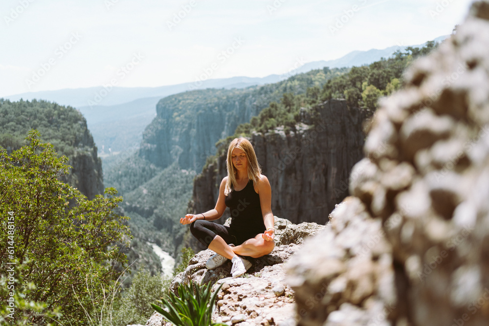 Naklejka premium Young beautiful girl woman in black sporty slim outfit meditating on edge rock mountain cliff peak with view on valley canyon. Picture of fit female hiker reached the mountain top.