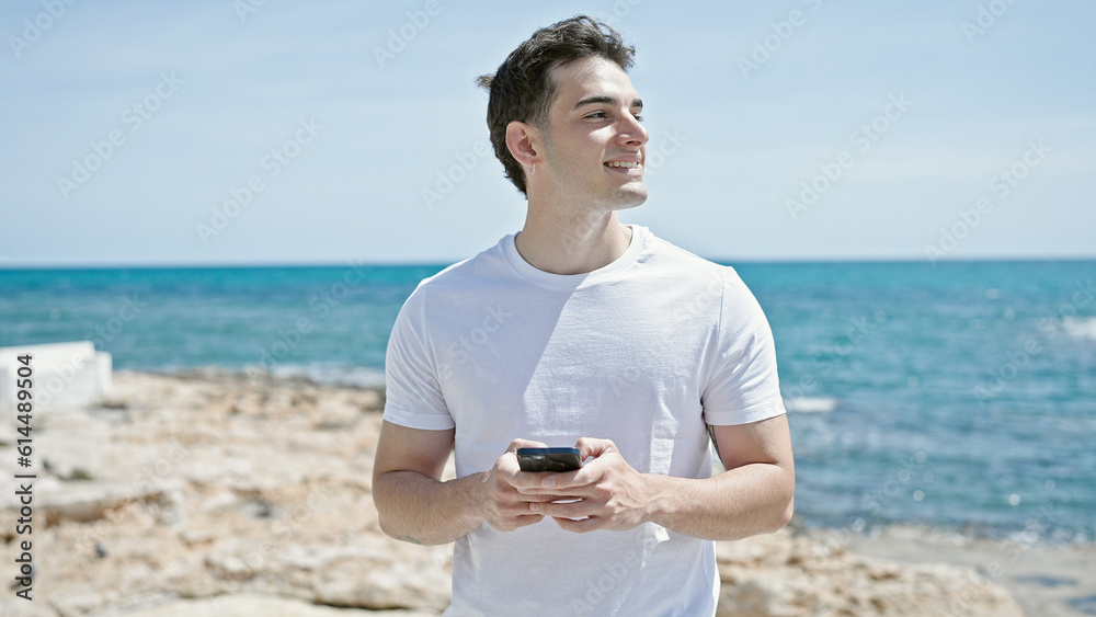 Young hispanic man smiling confident using smartphone at beach