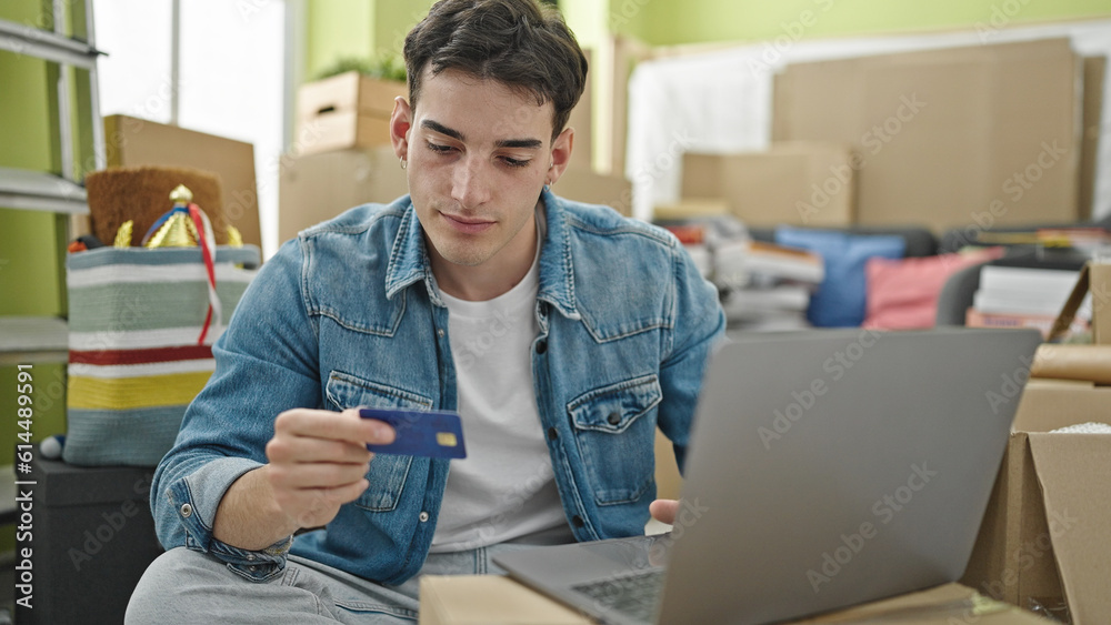 Young hispanic man using laptop and credit card sitting on sofa at new home