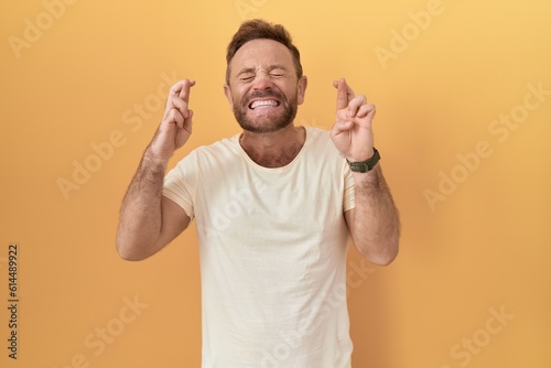 Middle age man with beard standing over yellow background gesturing finger crossed smiling with hope and eyes closed. luck and superstitious concept.