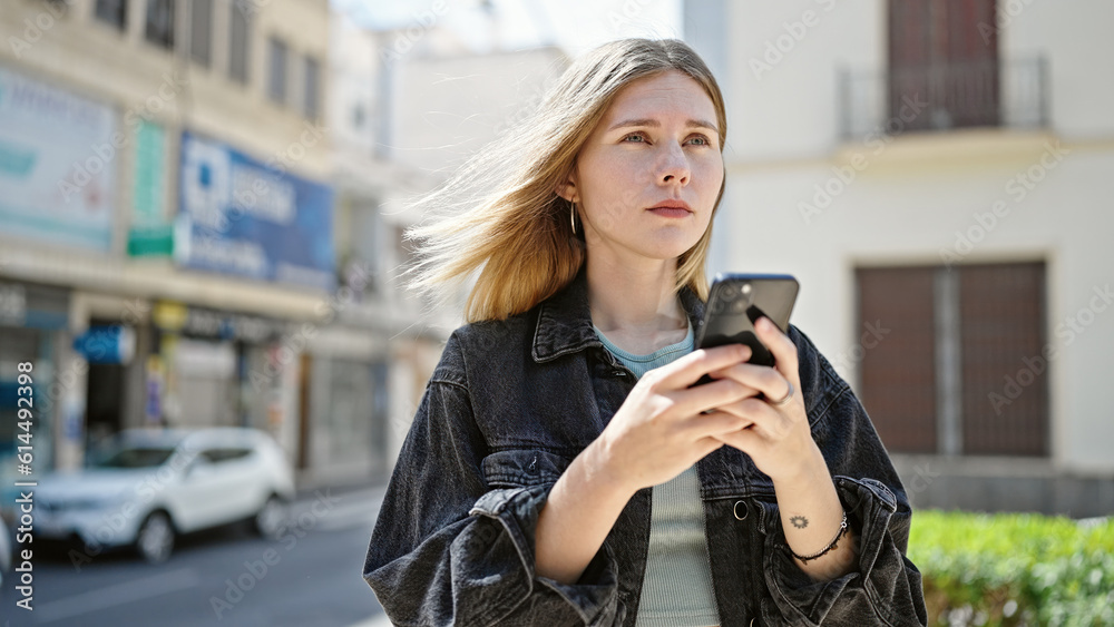 Young blonde woman using smartphone with serious face at street