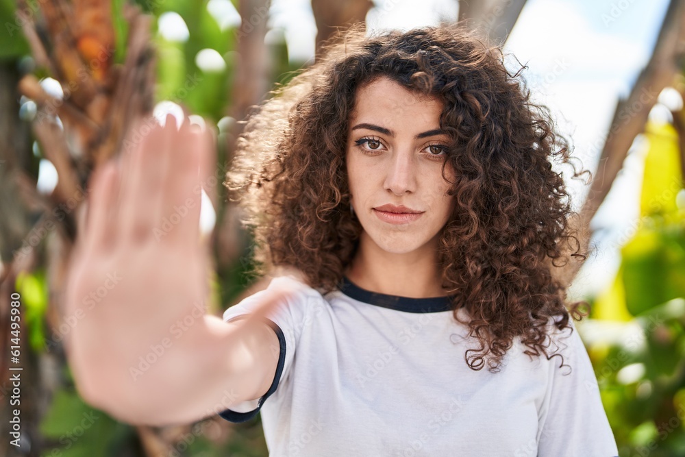 Fototapeta premium Hispanic woman with curly hair standing outdoors with open hand doing stop sign with serious and confident expression, defense gesture