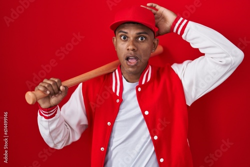 Wall Mural Young hispanic man playing baseball holding bat crazy and scared with hands on h