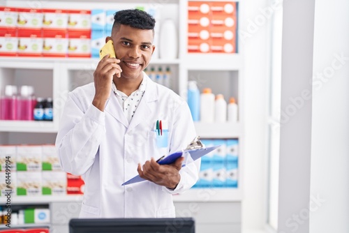 Young latin man pharmacist talking on smartphone reading report at pharmacy