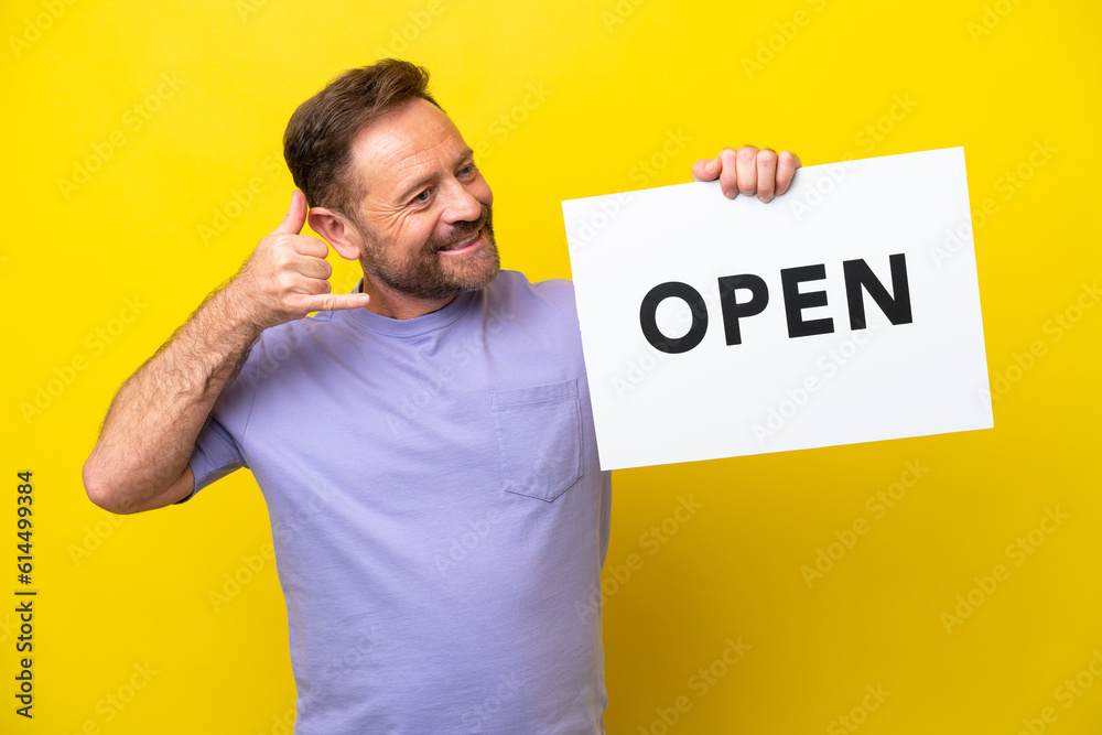 Middle age caucasian man isolated on yellow background holding a placard with text OPEN and doing phone gesture