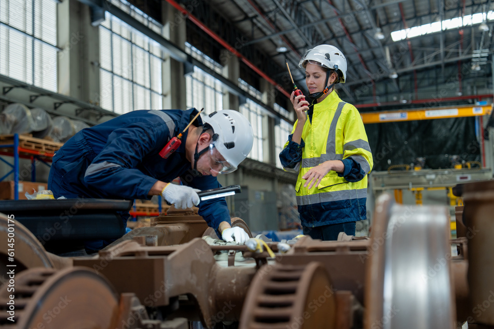 Female technician and engineer holding walkie talkie wearing helmet uniform inspects repair and maintenance of electric vehicle wheel and suspension system in electric train maintenance station.