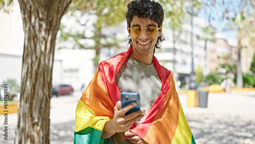 Young hispanic man wearing rainbow flag using smartphone at park