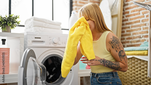 Young blonde woman washing clothes smelling dirty clothes at laundry room