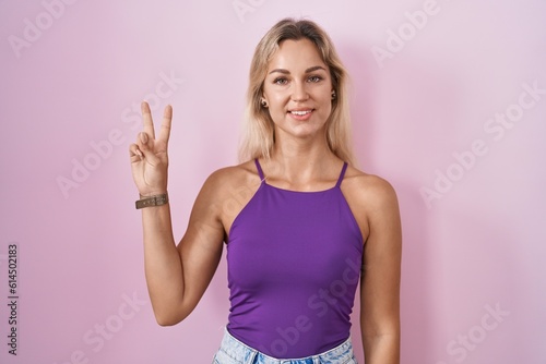 Young blonde woman standing over pink background showing and pointing up with fingers number two while smiling confident and happy.