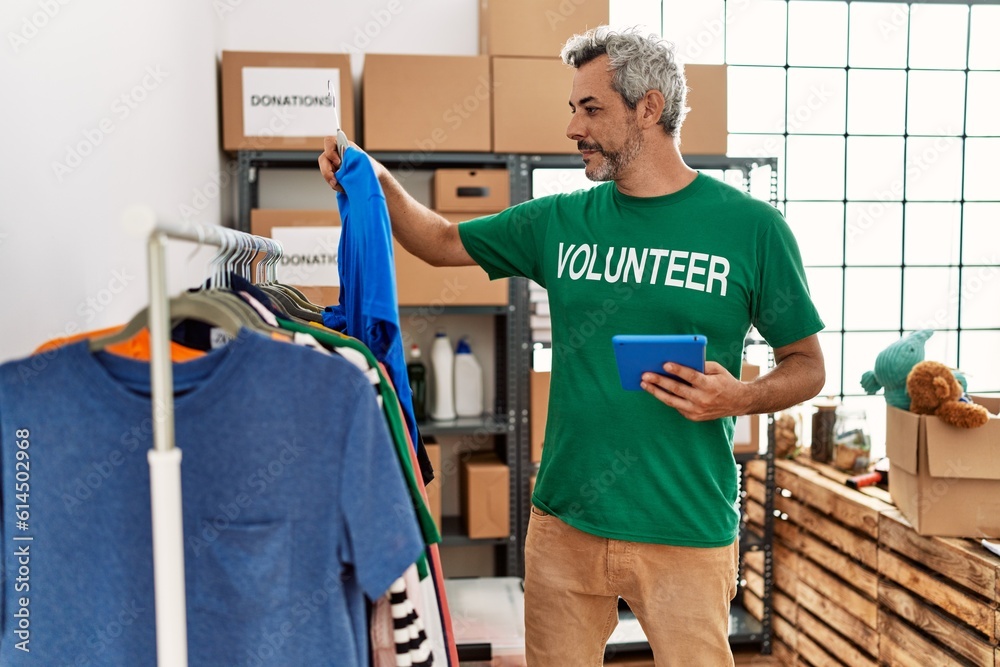 Middle age grey-haired man volunteer using touchpad holding clothes at charity center Middle age grey-haired man volunteer using touchpad holding clothes at charity center