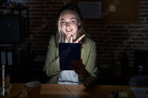 Blonde caucasian woman working at the office at night cheerful with a smile on face pointing with hand and finger up to the side with happy and natural expression