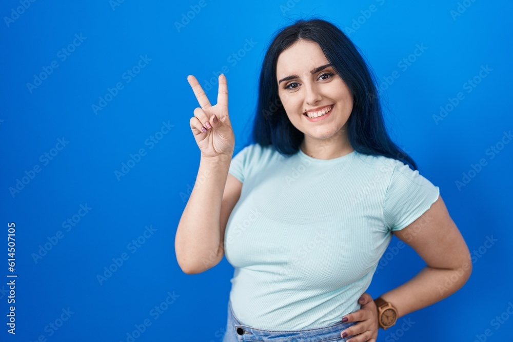 Fototapeta premium Young modern girl with blue hair standing over blue background smiling looking to the camera showing fingers doing victory sign. number two.