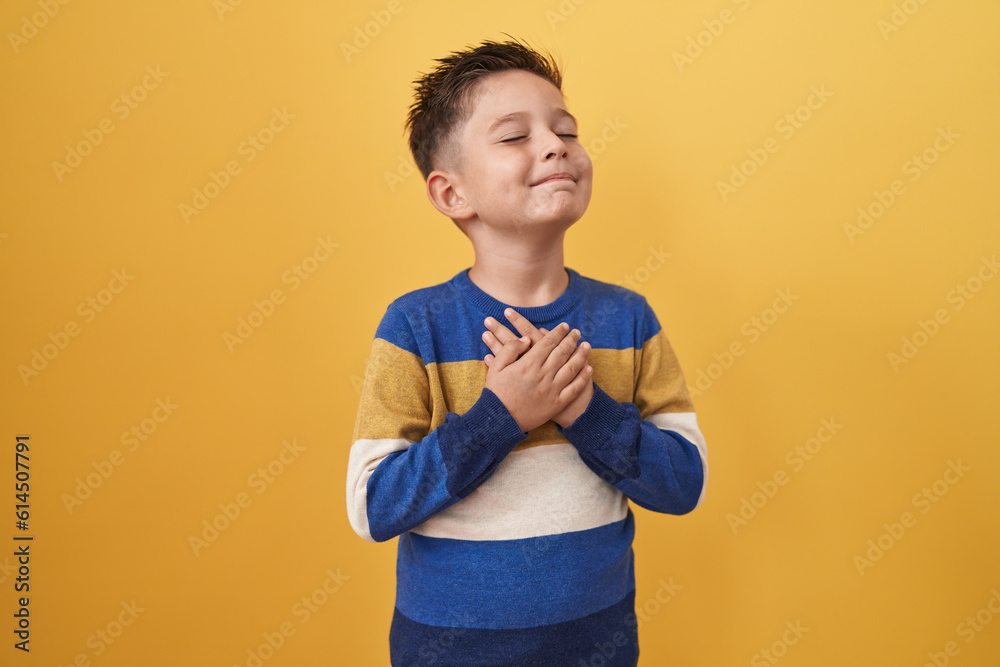 Little hispanic boy standing over yellow background smiling with hands on chest, eyes closed with grateful gesture on face. health concept. - obrazy, fototapety, plakaty 