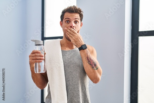 Young hispanic man wearing sportswear drinking water shocked covering mouth with hands for mistake. secret concept.