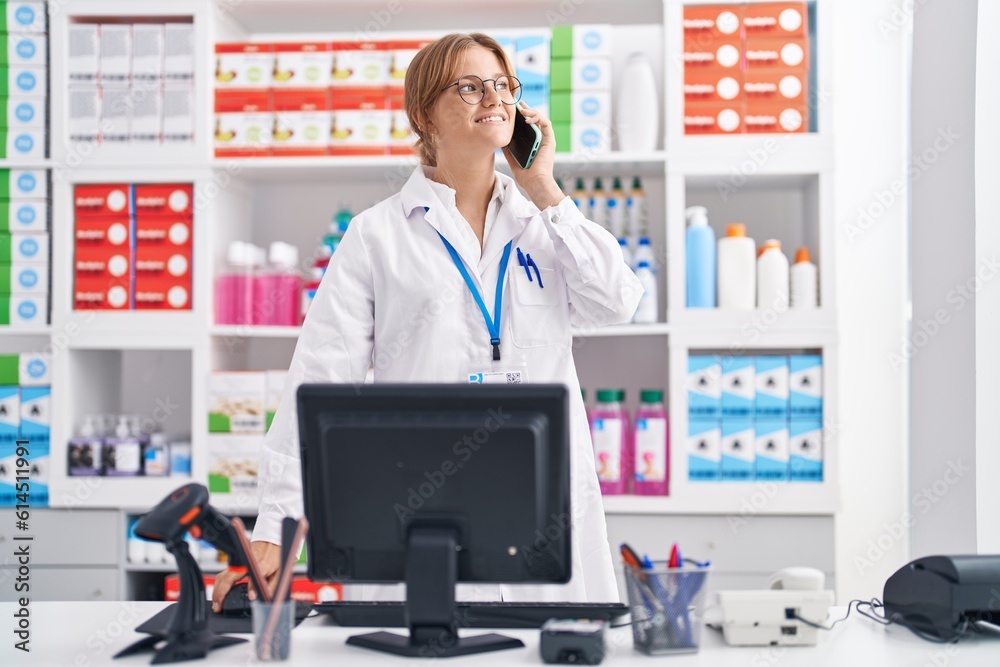 Young blonde girl pharmacist talking on smartphone using computer at pharmacy