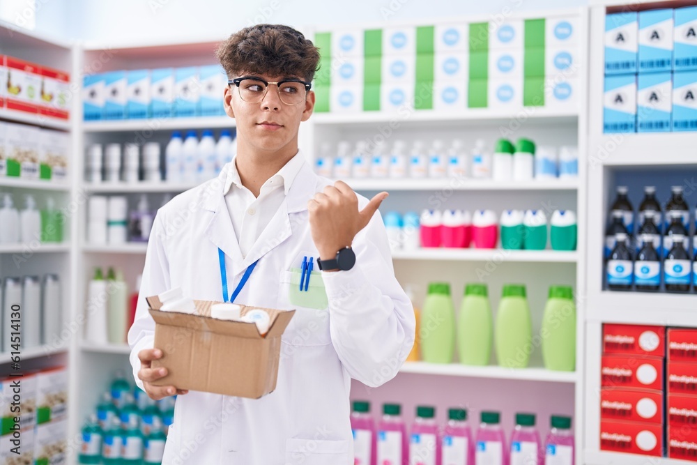 Hispanic teenager working at pharmacy drugstore holding box with pills pointing thumb up to the side smiling happy with open mouth
