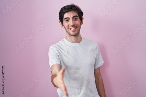 Young hispanic man standing over pink background smiling friendly offering handshake as greeting and welcoming. successful business.