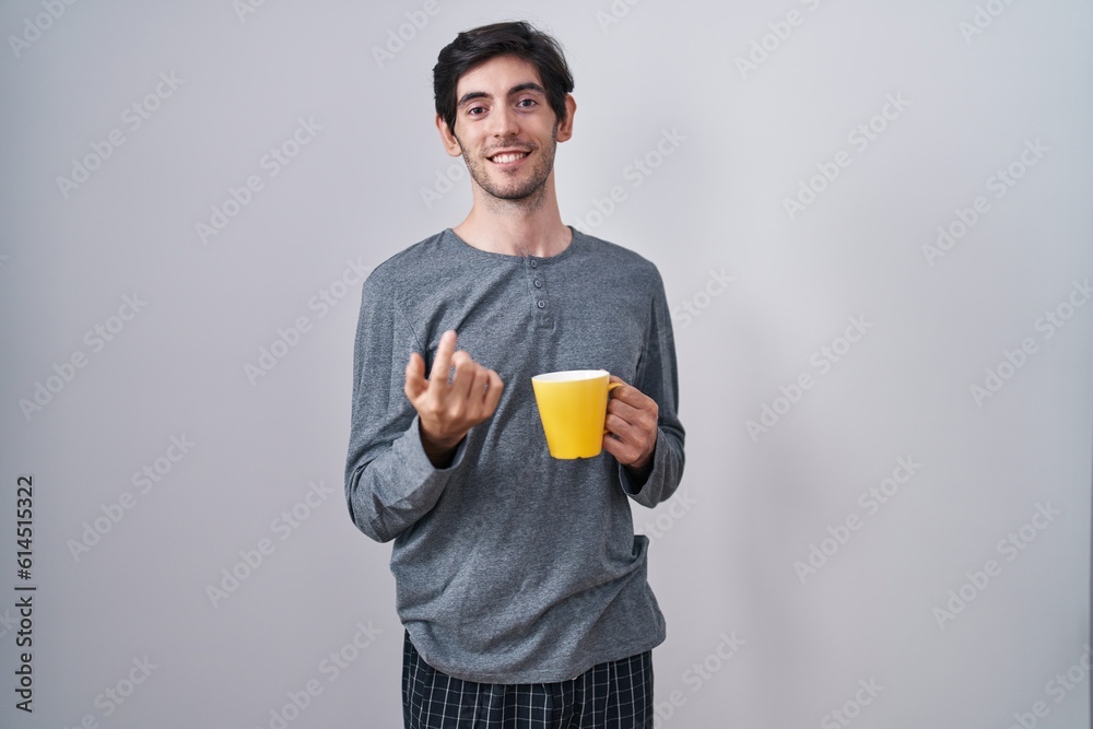 Young hispanic man wearing pajama drinking a cup of coffee beckoning come here gesture with hand inviting welcoming happy and smiling Young hispanic man wearing pajama drinking a cup of coffee beckoning come here gesture with hand inviting welcoming happy and smiling