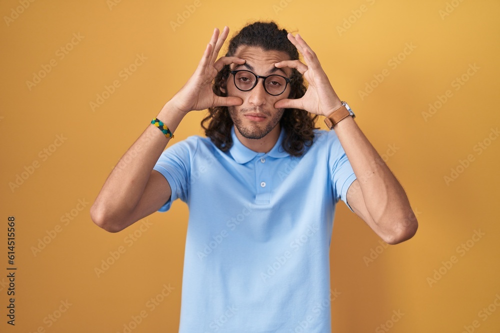 Young hispanic man standing over yellow background trying to open eyes with fingers, sleepy and tired for morning fatigue