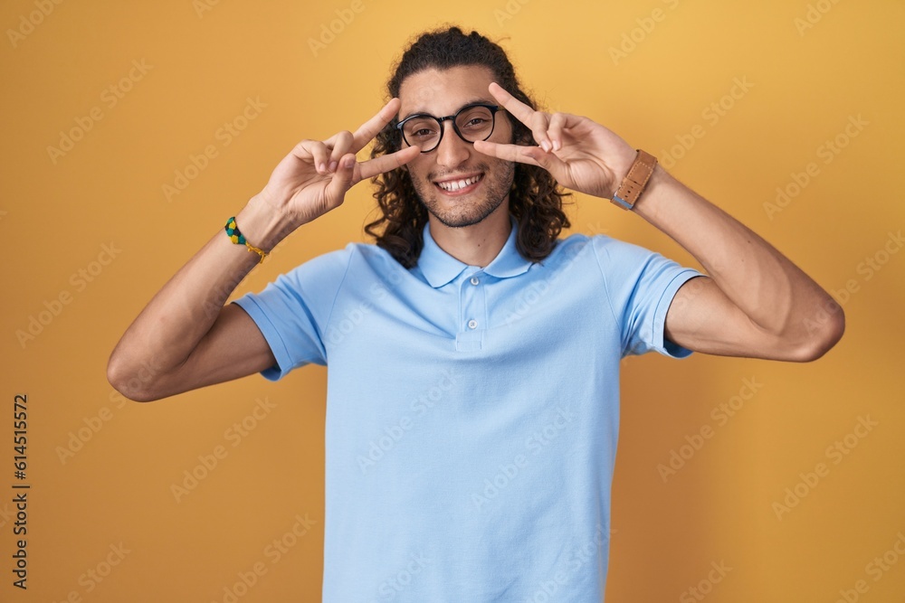Young hispanic man standing over yellow background doing peace symbol with fingers over face, smiling cheerful showing victory Young hispanic man standing over yellow background doing peace symbol with fingers over face, smiling cheerful showing victory