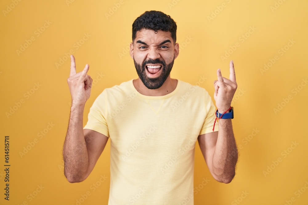 Hispanic man with beard standing over yellow background shouting with crazy expression doing rock symbol with hands up. music star. heavy music concept. Hispanic man with beard standing over yellow background shouting with crazy expression doing rock symbol with hands up. music star. heavy music concept.