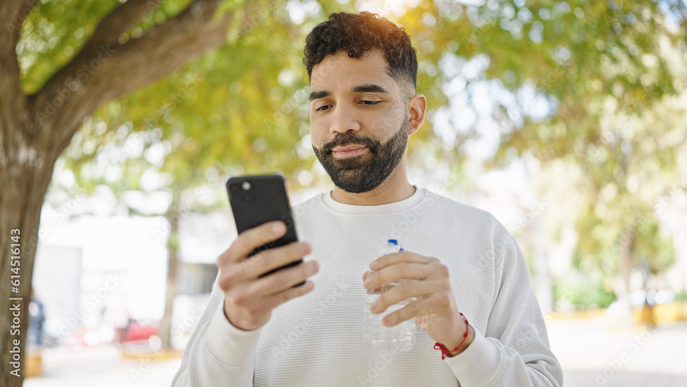 Young hispanic man holding bottle of water using smartphone at park Young hispanic man holding bottle of water using smartphone at park
