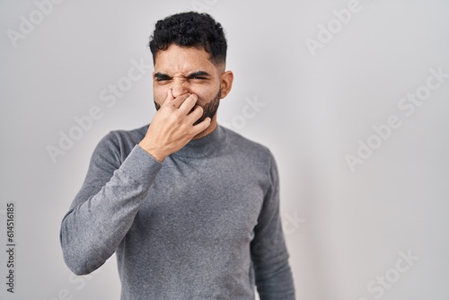 Hispanic man with beard standing over white background smelling something stinky and disgusting, intolerable smell, holding breath with fingers on nose. bad smell