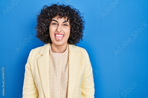 Young brunette woman with curly hair standing over blue background sticking tongue out happy with funny expression. emotion concept.