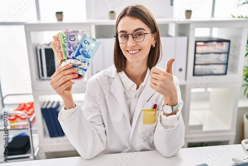 Young caucasian doctor woman holding canadian dollars banknotes smiling happy and positive  thumb up doing excellent and approval sign