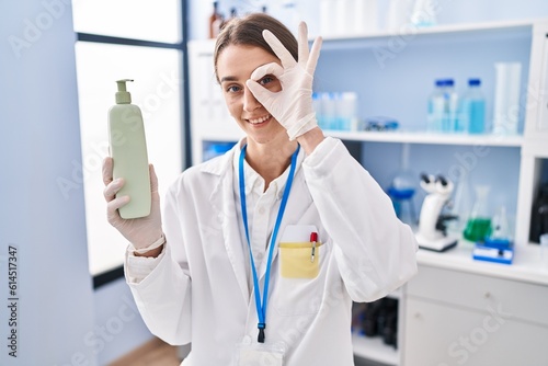 Young caucasian woman working at scientist laboratory holding body lotion smiling happy doing ok sign with hand on eye looking through fingers