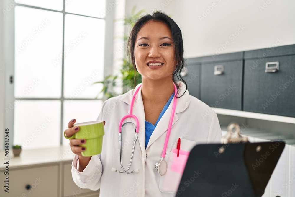 Young chinese woman wearing doctor uniform holding clipboard drinking coffee at clinic Young chinese woman wearing doctor uniform holding clipboard drinking coffee at clinic