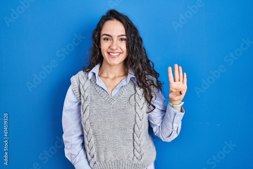 Young brunette woman standing over blue background showing and pointing up with fingers number four while smiling confident and happy.